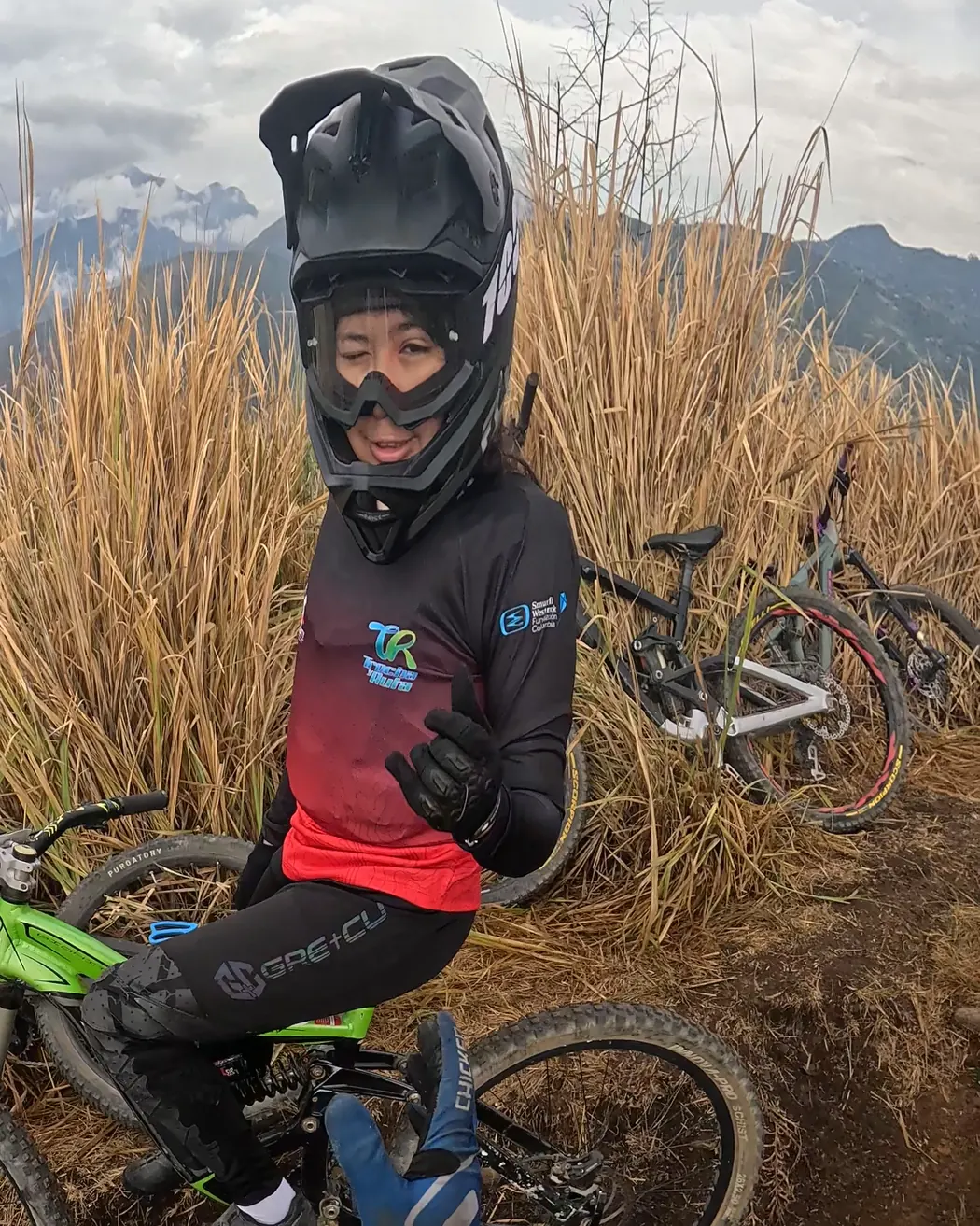 Isabel Quiñones del Club Trocha y Ruta, con casco integral y gafas, guiña un ojo y levanta el pulgar sentada en la bicicleta verde, entre pasto seco y montañas, en lo alto del Cali Bike Park, 2026
