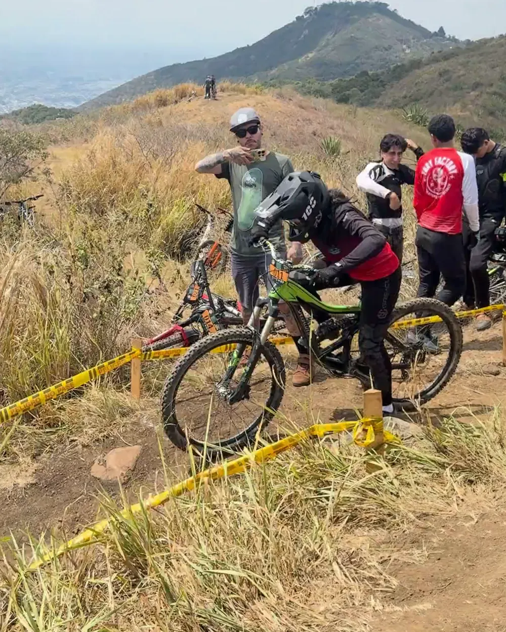Isabel Quiñones del Club Trocha y Ruta, con casco integral, sobre la bicicleta de descenso verde en la zona de salida, rodeada de otros corredores, Copa Cali Bike Park, 2026