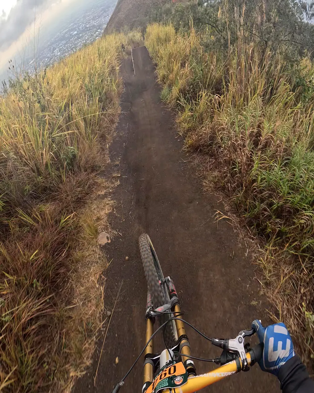 Vista desde el pecho de un corredor, sobre el manubrio: una recta de tierra entre pasto alto va hacia un cerro, Cali Bike Park, 2026