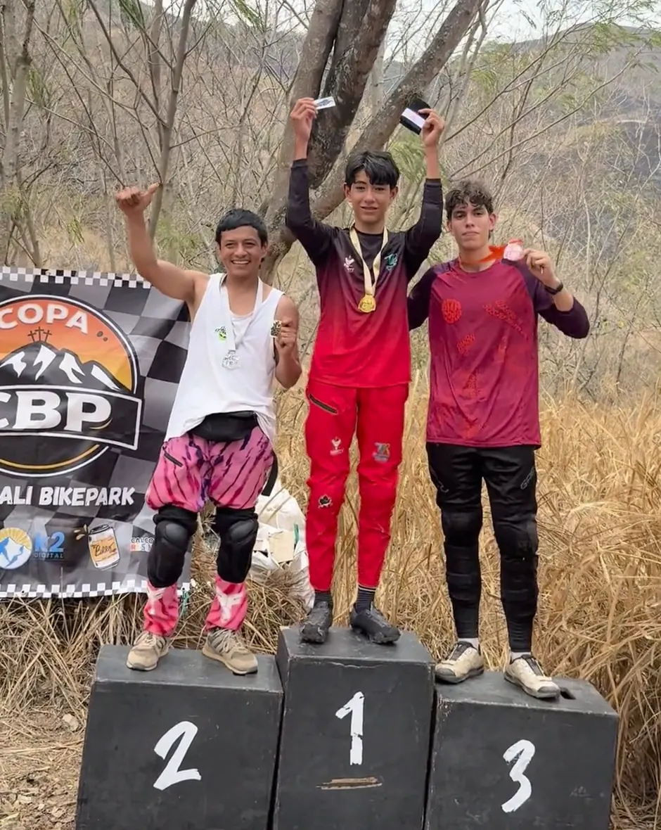 Podio de la Copa Cali Bike Park: Samuel Ortiz del Club Trocha y Ruta en el cajón 1, con la medalla puesta y los brazos arriba, junto a los corredores de los cajones 2 y 3, frente al pendón de la copa, 2026