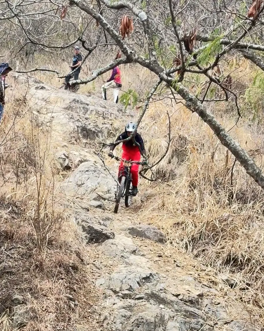 Samuel Ortiz del Club Trocha y Ruta baja por una placa de roca entre pasto seco y ramas, con espectadores arriba, Copa Cali Bike Park, 2026