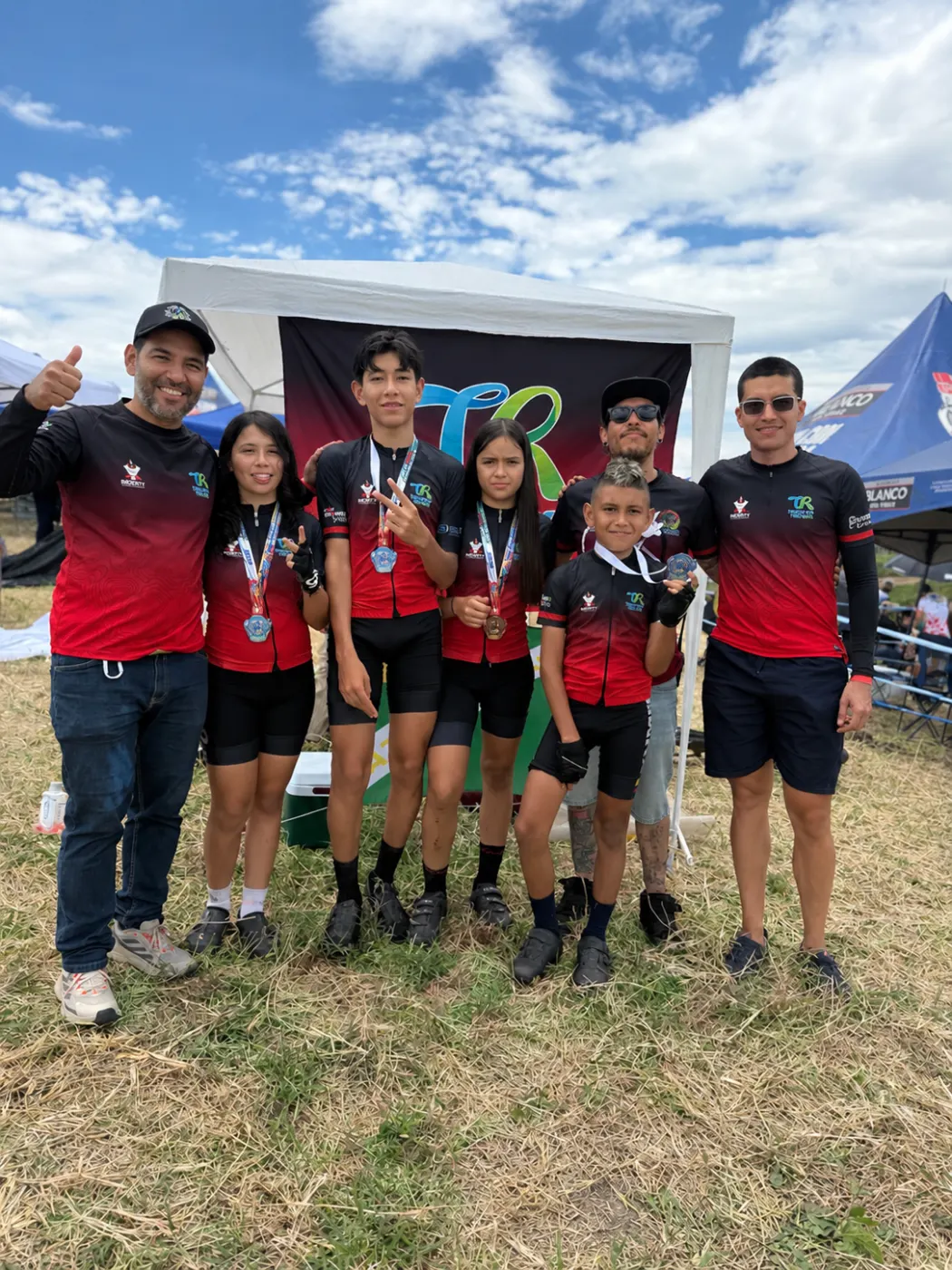 Los cuatro corredores premiados del Club Trocha y Ruta, con sus medallas, junto a tres acompañantes bajo la carpa del club con la bandera de Trocha y Ruta de fondo, en la Copa Let's Go Interdepartamental XCO, Alcalá, 2026