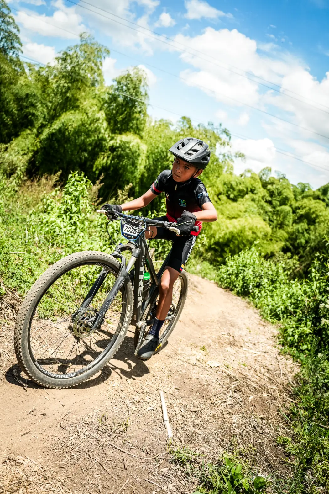 Jostin Villamizar del Club Trocha y Ruta, dorsal 7026, sube un sendero angosto entre monte en el circuito de la Copa Let's Go Interdepartamental XCO, Alcalá, 2026